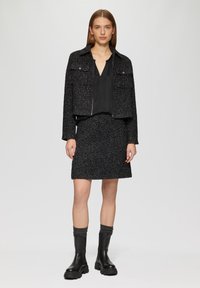 Black tweed jacket with two front pockets, paired with a black blouse and a matching skirt. Black knee-high boots with a thick sole.