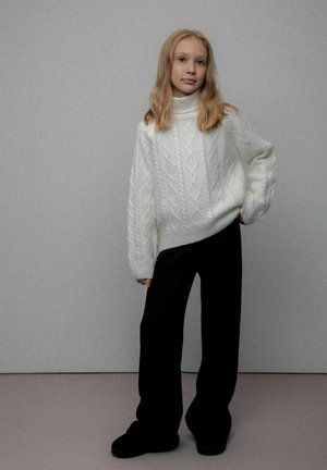 Młoda osoba z długimi blond włosami, ubrana w biały sweter z golfem z splotem warkoczowym i luźne czarne spodnie, stojąca na jednolitym tle.