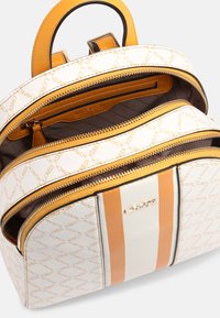Sac à dos avec un extérieur texturé blanc, présentant des accents orange, plusieurs compartiments zippés et un design de logo imprimé.