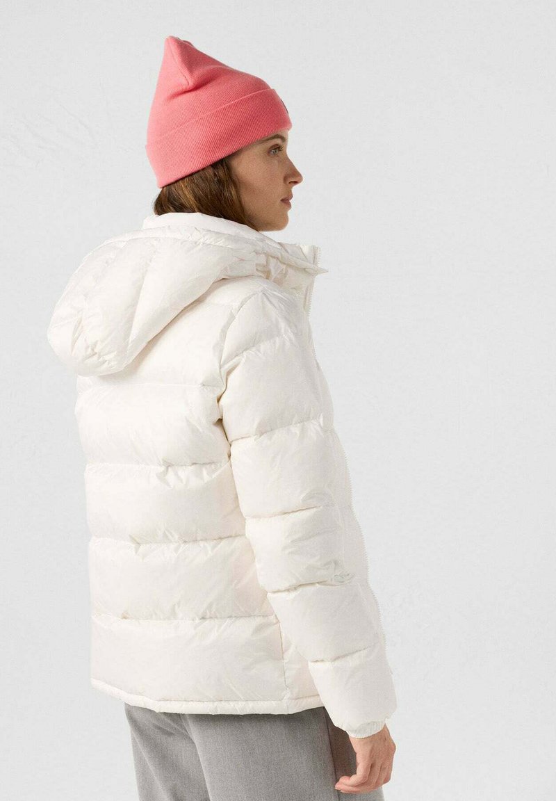 Witte donzen jas met capuchon, doorgestikt ontwerp en geribde boorden. Gecombineerd met een lichtroze muts. Model van opzij gezien.