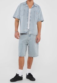 Man in een lichtblauwe denim korte mouwen shirt en bijpassende shorts, witte tanktop, witte sokken en zwarte sneakers, staand tegen een witte achtergrond.