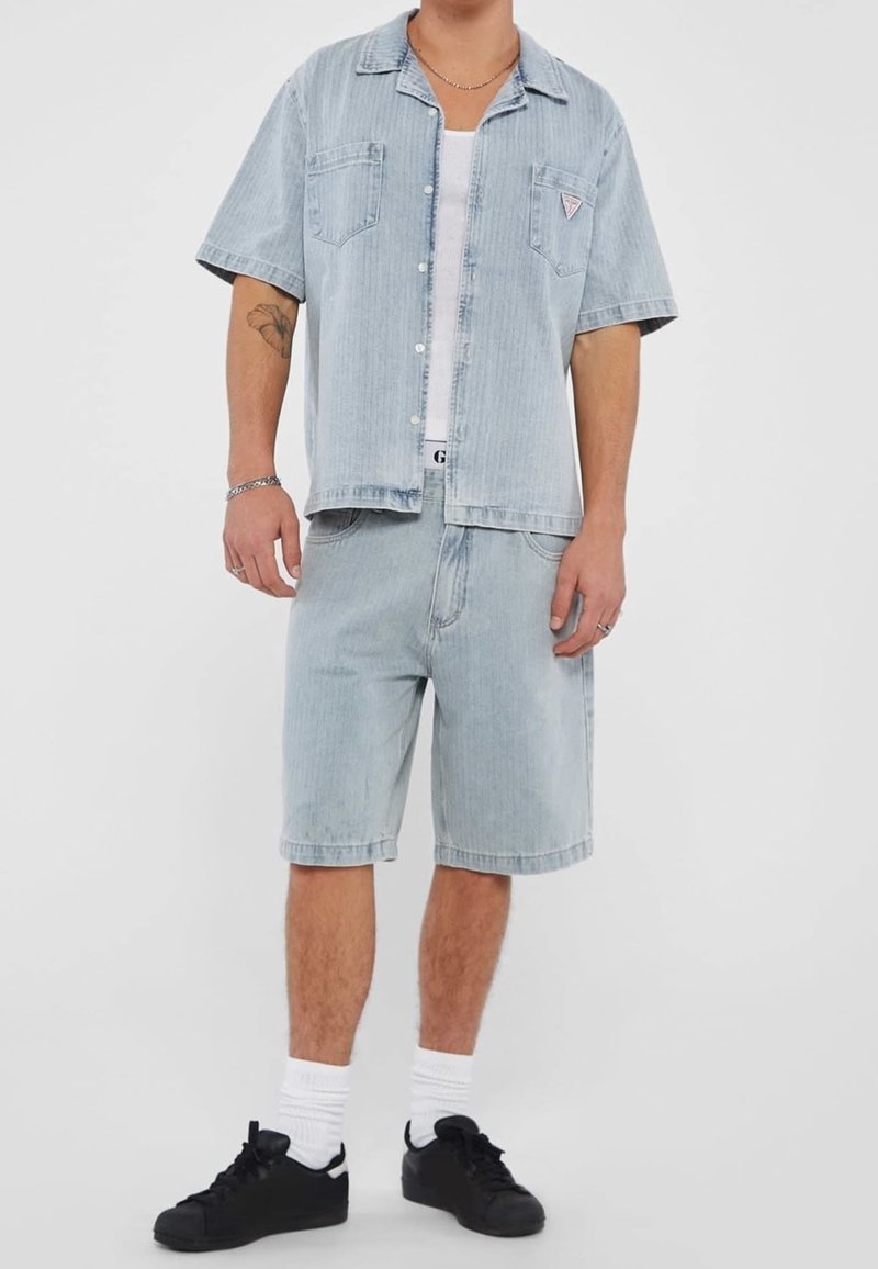 Man in een lichtblauwe denim korte mouwen shirt en bijpassende shorts, witte tanktop, witte sokken en zwarte sneakers, staand tegen een witte achtergrond.
