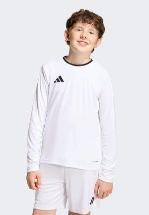 Lächelnder Junge mit lockigem Haar, der ein weißes Adidas Langarmshirt und passende Shorts trägt, steht vor einem schlichten hellen Hintergrund.