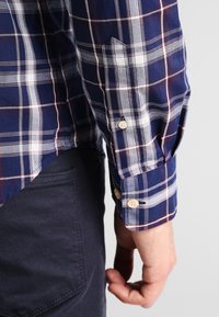 Niebieska koszula w kratę z białymi i czerwonymi akcentami, z mankietami na guziki i zaokrąglonym dołem, wykonana z lekkiego materiału.