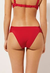 Červené bikiny s řasením na bocích, vyrobené z hladkého materiálu, s mírně zvýšeným pasem a minimalistickým střihem.