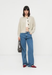 Strikket beige cardigan med brune knapper,nå med høyt liv blå jeans og røde sko. Holder en liten brun håndveske.