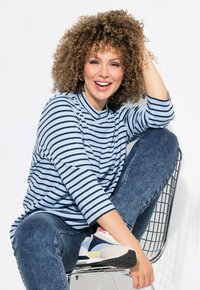 Randig ljusblå långärmad topp, mörka denimjeans och flerfärgade sneakers; sittande på en trådstol med vit bakgrund.