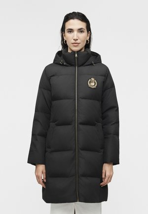 Nő hosszú fekete kabátot visel, kapucnival és arany emblémával a mellkasán, egyszínű világos háttér előtt áll.