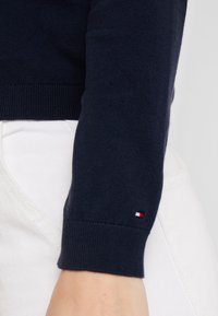 Navyblauer gestrickter Pullover mit gerippten Bündchen und einem kleinen Tommy Hilfiger-Logo in Rot, Weiß und Blau am Handgelenk.