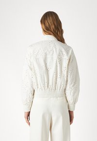 Kvinna med långt hår står vänd bort, iförd en vit ögla spetss bomberjacka och högmidjade vita byxor.