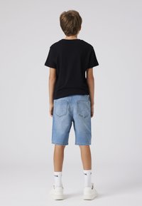 Črna bombažna majica, kratke kavbojke svetlo modre barve, pogled od zadaj. Kratke hlače imajo zavihane robove in dva zadnja žepa. Bele superge s srednje dolgimi nogavicami.