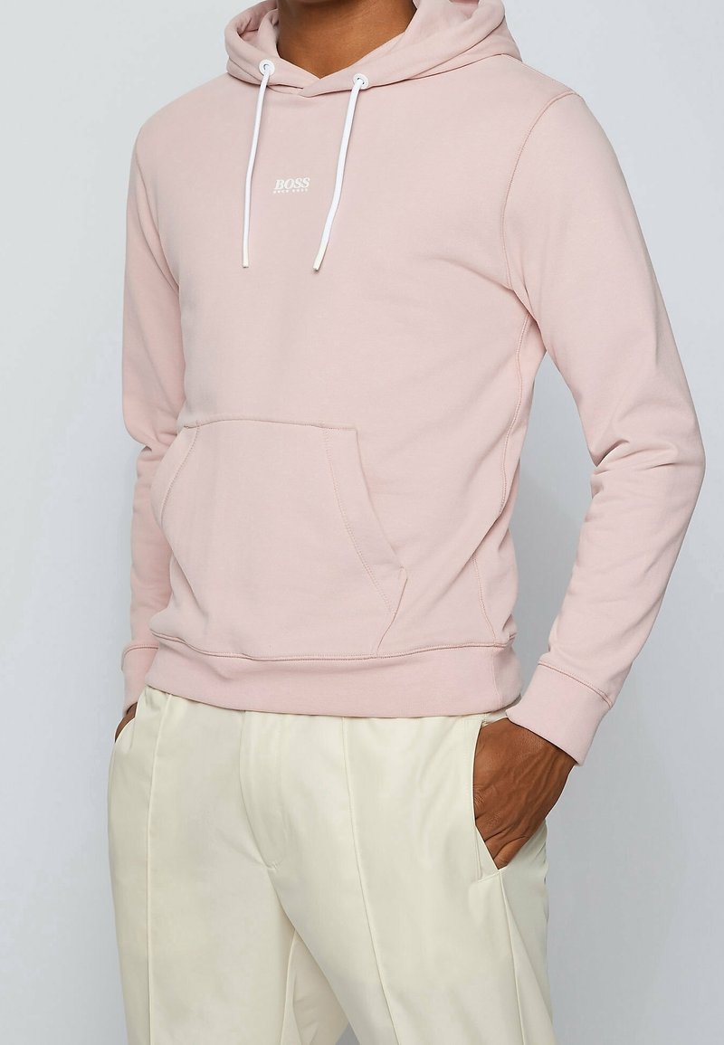 Personne portant un sweat à capuche BOSS rose clair avec poche avant et cordons blancs, associé à un pantalon couleur crème, mains dans les poches.