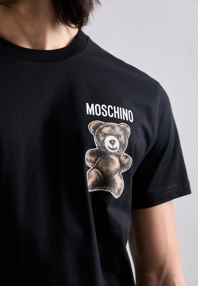 Black cotton t-shirt featuring a graphic of a teddy bear on the left chest with "MOSCHINO" text above. Short sleeves and crew neck design.