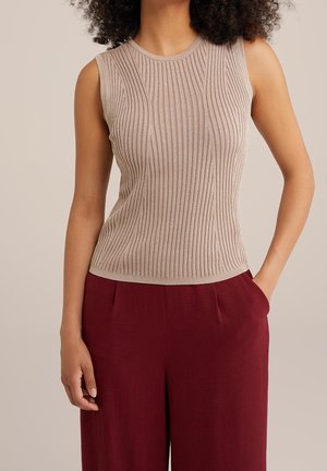 Femme portant un haut beige côtelé sans manches rentré dans un pantalon bordeaux taille haute, une main dans la poche.