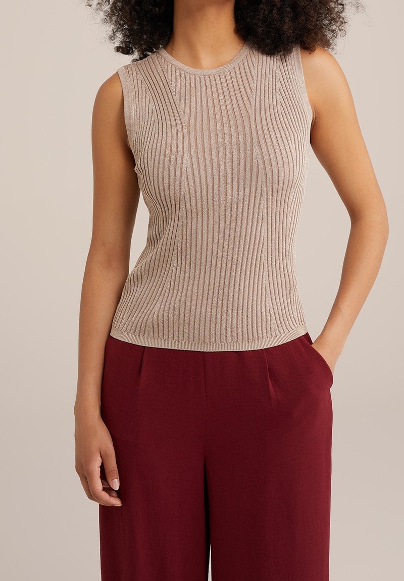 Femme portant un haut beige côtelé sans manches rentré dans un pantalon bordeaux taille haute, une main dans la poche.