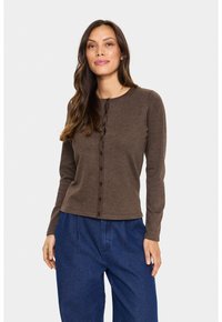 Ruskea pitkähihainen neuletakki, jossa on pyöreä kaula-aukko ja nappikiinnitys edessä. Pehmeä rakenne ja istuva muotoilu, yhdistettynäsinisiin leveälahkeisiin housuihin.