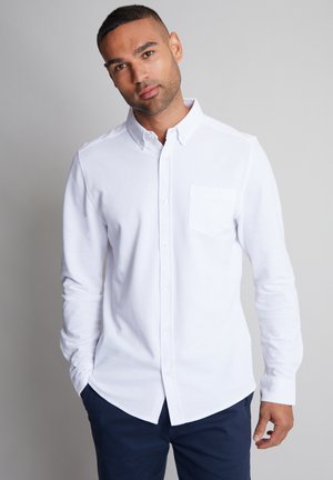 Camisa blanca de manga larga, con botones y cuello corto, un bolsillo en el pecho y dobladillo redondeado, combinada con pantalones oscuros. Textura suave.