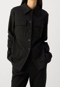Czarna zapinana na guziki kurtka z kołnierzem, z dwoma kieszeniami na piersi i gładkim, teksturowanym materiałem. Zaprojektowana z luźnym krojem.