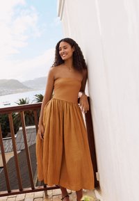 Pánt nélküli, mustárszínű ruha redős szoknyával, könnyű anyagból. A modell egy fehér falnak támaszkodik, bemutatva a ruha dizájnját.