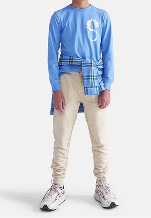 Młoda osoba ubrana w niebieską koszulę z długim rękawem, beżowe dresowe spodnie, białe tenisówki, z niebieską koszulą w kratę zawiązaną w talii.