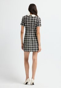 Donna che indossa un vestito a maniche corte a scacchi bianco e nero con colletto bianco, in piedi, di spalle, e indossa scarpe con tacco alto bianche.