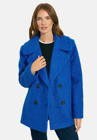 Sininen kaksirivinen villatakki, jossa on suuret kaulukset, teksturoitu pinta, neljä nappia ja rento istuvuus. Päällä musta paita.