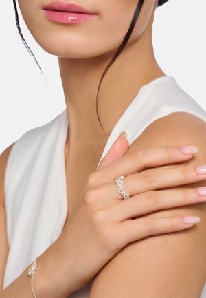 Frau trägt ein weißes ärmelloses Oberteil und zeigt einen Ring mit Diamantblumenmuster an ihrem Finger sowie ein passendes Armband an ihrem Handgelenk.