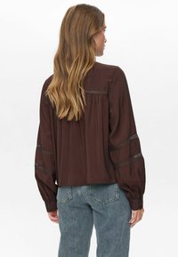 Vrouw met lang golvend haar, gekleed in een losse donkerbruine blouse met kantdetails op de mouwen en rug, gecombineerd met high-waisted blauwe jeans.