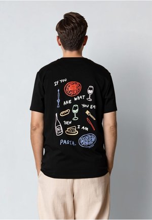Person som har på seg en svart t-skjorte med fargerike pasta-tema illustrasjoner og tekst som lyder "Hvis du er det du spiser, så er jeg pasta."