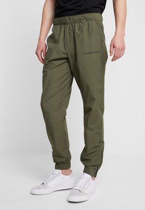 Homme portant un jean Calvin Klein vert olive avec une ceinture et des poignets élastiques, associé à des baskets blanches sur un sol gris.