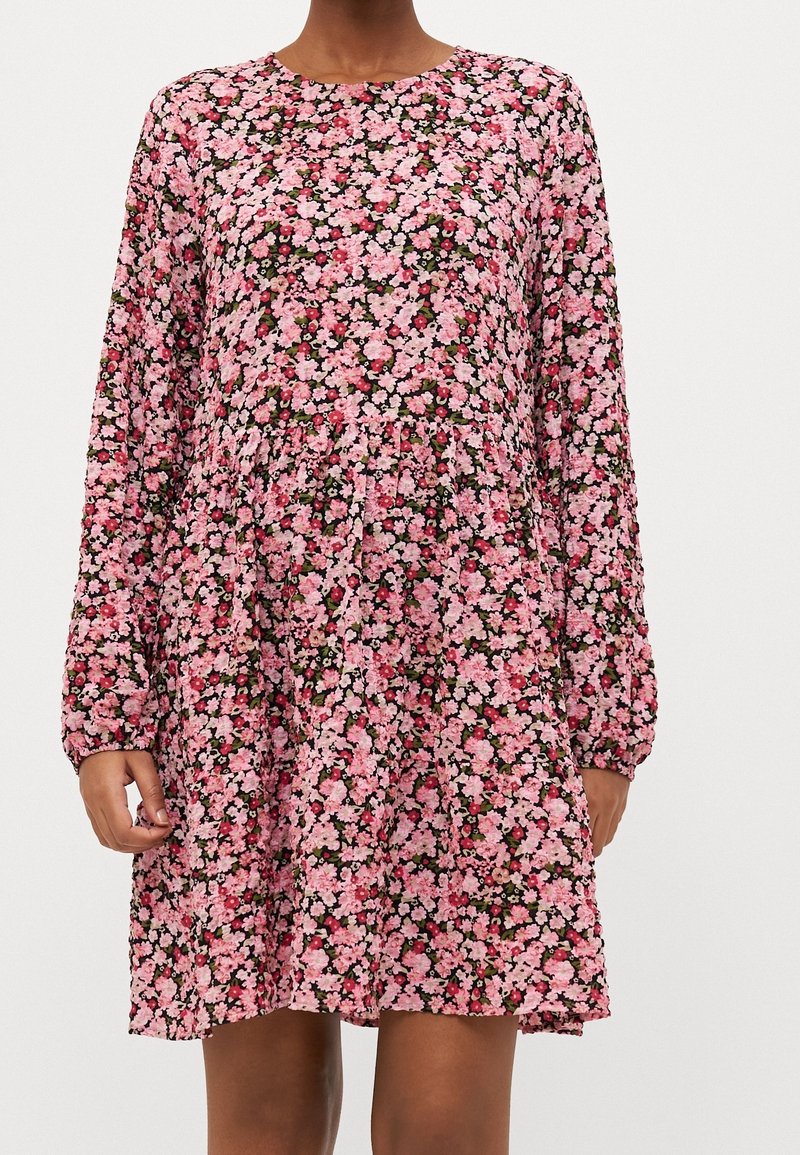 Femme portant une robe à manches longues, longueur genou, avec un motif floral rose dense sur un fond sombre, debout devant un fond uni.