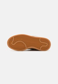 Suela de zapatilla con un patrón de espiga, hecha de un material de goma marrón y con una superficie texturizada para mayor tracción. Parte superior blanca parcialmente visible.
