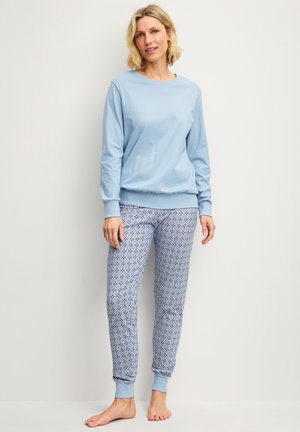 Vrouw staat op blote voeten, draagt een lichtblauw shirt met lange mouwen en een blauw-witte patroon pyjamabroek tegen een effen achtergrond.