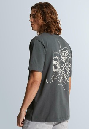 Młody mężczyzna z włosami do ramion, noszący ciemnoszary T-shirt z dużym białym abstrakcyjnym kwiatowym wzorem na plecach.
