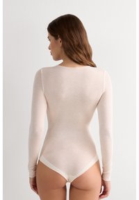 Body à manches longues en beige clair, fabriqué en tissu doux. Il présente un col arrondi et un design sans coutures, avec une coupe string à la base.