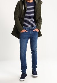 Parka verde oliva con capucha, camiseta a rayas en azul marino y blanco, jeans de mezclilla azules y zapatillas oscuras. Presenta un diseño casual.