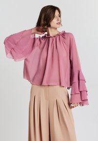 Donna che indossa un leggero blouse mauve con maniche a campana a strati e pantaloni beigi a vita alta con pieghe, in posa con la mano dietro al collo.