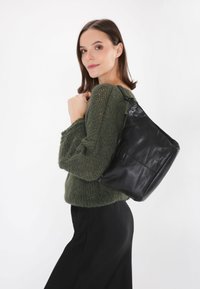 Černá kožená kabelka s hladkým povrchem, volně tvarovaná, s přední kapsou na zip, nošena v ležérním stylu přes zelený pletený svetr.