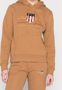 Felpa brown con tasca frontale, logo ricamato e cappuccio con lacci. Abbinata a pantaloni brown coordinati con un piccolo logo.