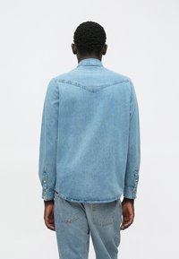 Ανοιχτό μπλε τζιν πουκάμισο με κομμένο γιακά, κουμπωτές μανσέτες και καμπυλωτό στρίφωμα. Διαθέτει σχέδιο γιόκ στο πίσω μέρος.