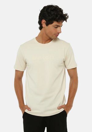 Uomo con maglietta a maniche corte color crema con testo in rilievo sottile, mani nelle tasche, guarda verso il basso e a destra, in piedi su sfondo bianco.