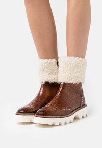 Botas de tobillo de cuero marrón con un patrón texturizado, puños de borreguillo blanco, acentos de tachuelas plateadas y una suela gruesa blanca.