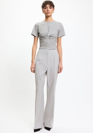 Kobieta ubrana w jasno szary, krótki rękaw z węzłem, zestawiona z wysokim stanem, szerokimi szarymi spodniami i czarnymi spiczastymi butami, stojąca prosto.