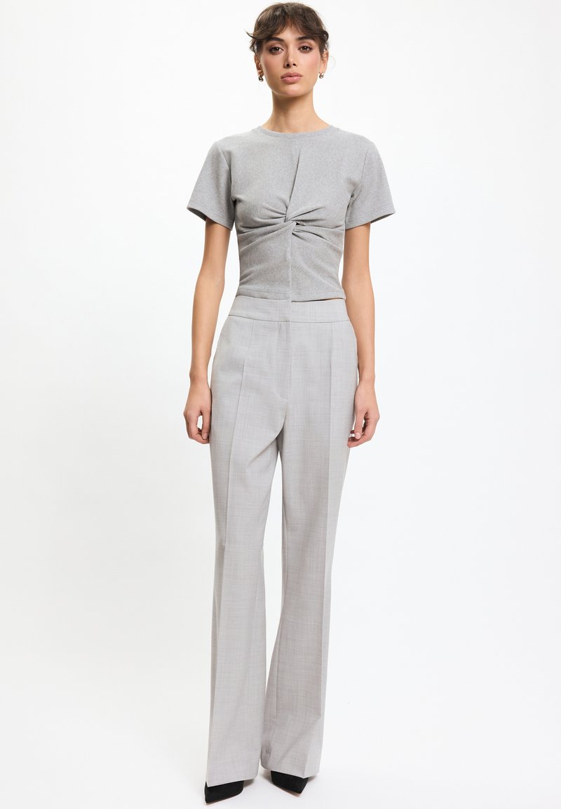 Vrouw draagt een lichtgrijs kort mouw top met een gedraaide knoop, gecombineerd met een hooggetailleerde, wijde grijze broek en zwarte puntige schoenen, staat rechtop.