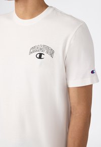 Camiseta de manga corta blanca con el texto y el logo en negro de "Champion" en el pecho y un pequeño logo multicolor de Champion en la manga izquierda.
