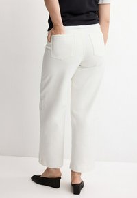 Person trägt weiße Hosen mit weitem Bein und aufgesetzten Gesäßtaschen, schwarzes Oberteil und schwarze, flache Slipper. Steht mit Händen in den Taschen.