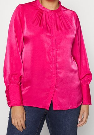 Kobieta ubrana w jasnoróżową satynową bluzkę z długimi bufkami i zatrzaskami, w połączeniu z ciemnoniebieskimi dżinsami, na białym tle.