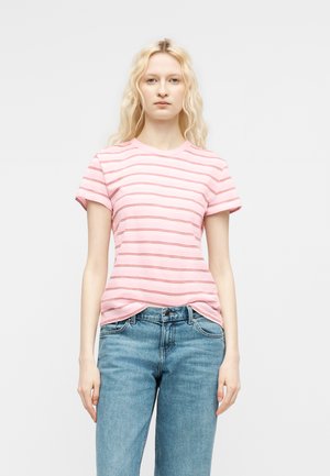 Młoda kobieta o długich blond włosach, ubrana w różowy, krótkim rękawem, paskowany T-shirt i niebieskie dżinsy, stojąca na jednolitym białym tle.