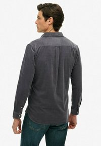 Man in een lange donkergrijze corduroy overhemd met lange mouwen en een blauwe spijkerbroek, gezien van achteren tegen een witte achtergrond.