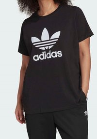 Osoba oblečená v černém tričku Adidas s bílým logem trefoil a shodnými černými kalhotami, ruka v kapse, na jednoduchém světlém pozadí.